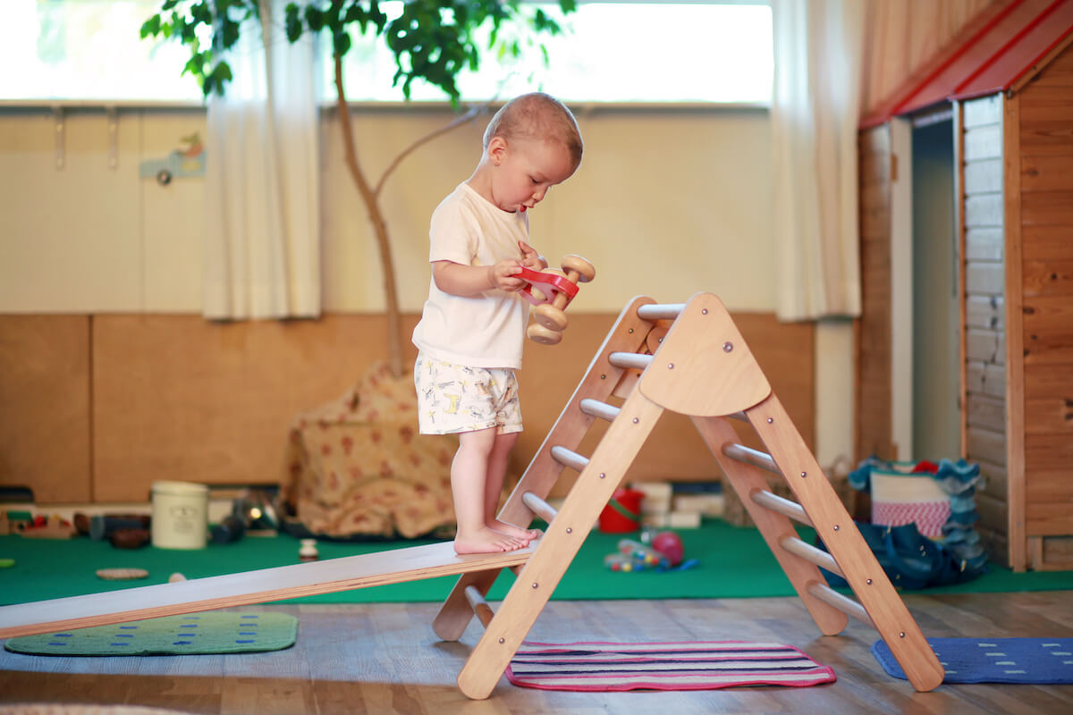 Montessori playroom: child standing on a Pikler triangle while holding a wooden car